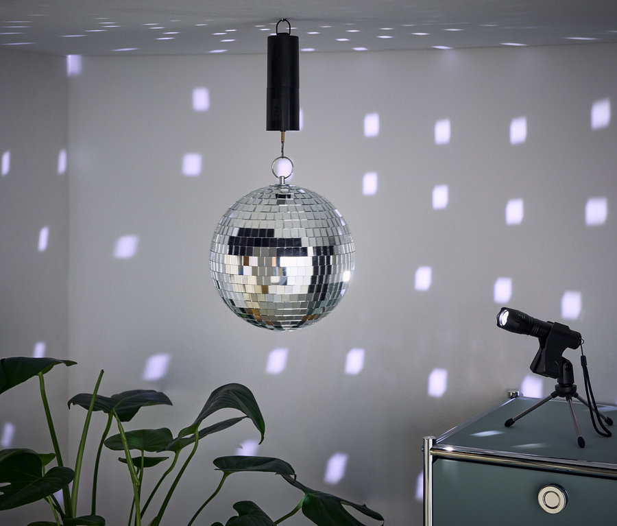 Eine Spiegelkugel hängt von der Decke und wirft Lichtflecken an die Wände. Daneben steht ein Scheinwerfer auf einem Schrank und im Vordergrund ist eine Pflanze.