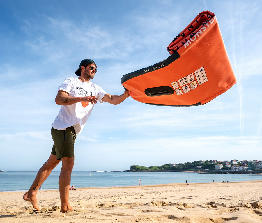 Mann hält ein zusammengerolltes oranges Aqua Marina SUP Board »Monster« 12'0" am Strand.