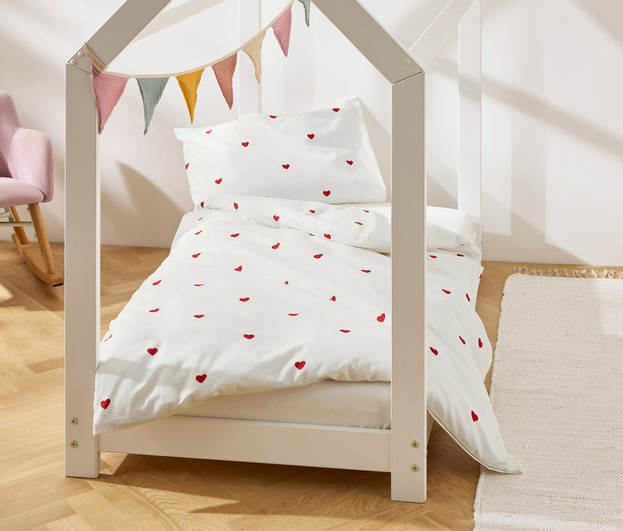 Ein Kinderbett in Hausform mit weißer Bettwäsche mit roten Herzen und Wimpeln. Im Hintergrund ein rosa Sessel.