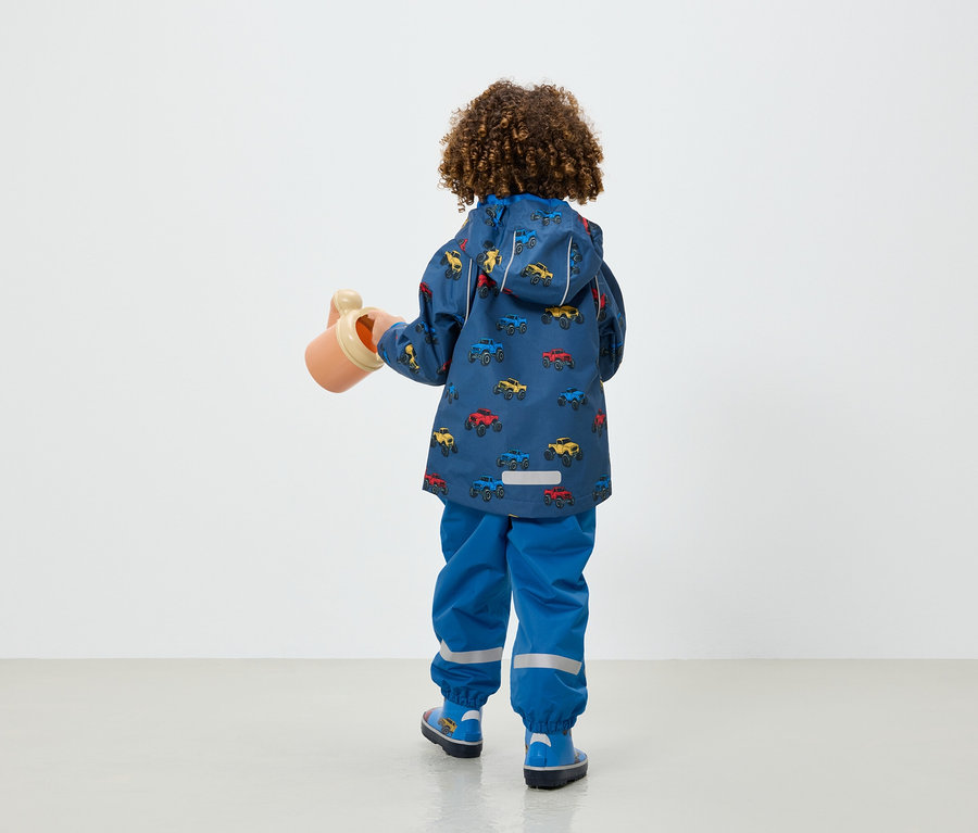 Junge in blauer Jacke und Hose mit Autoaufdruck hält eine beige Gießkanne.