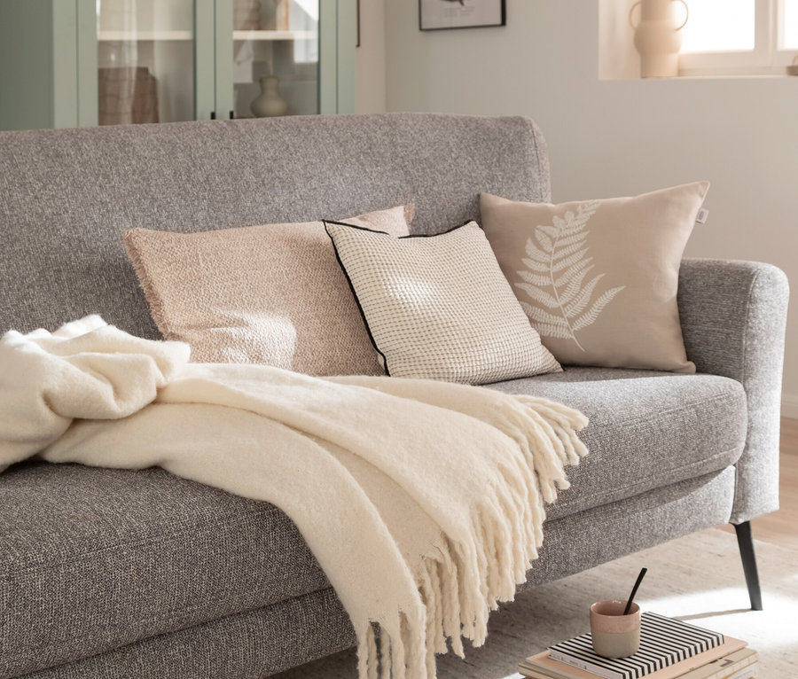 Graues Sofa mit drei Kissen und einer cremefarbenen Decke. Auf dem Boden liegt ein Stapel Bücher mit einer Tasse.