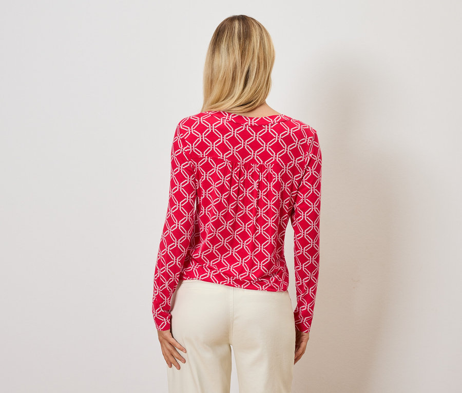 Von hinten ist eine Frau mit blonden Haaren zu sehen, die ein rotes Oberteil mit einem weißen geometrischen Muster und cremefarbene Hosen trägt.