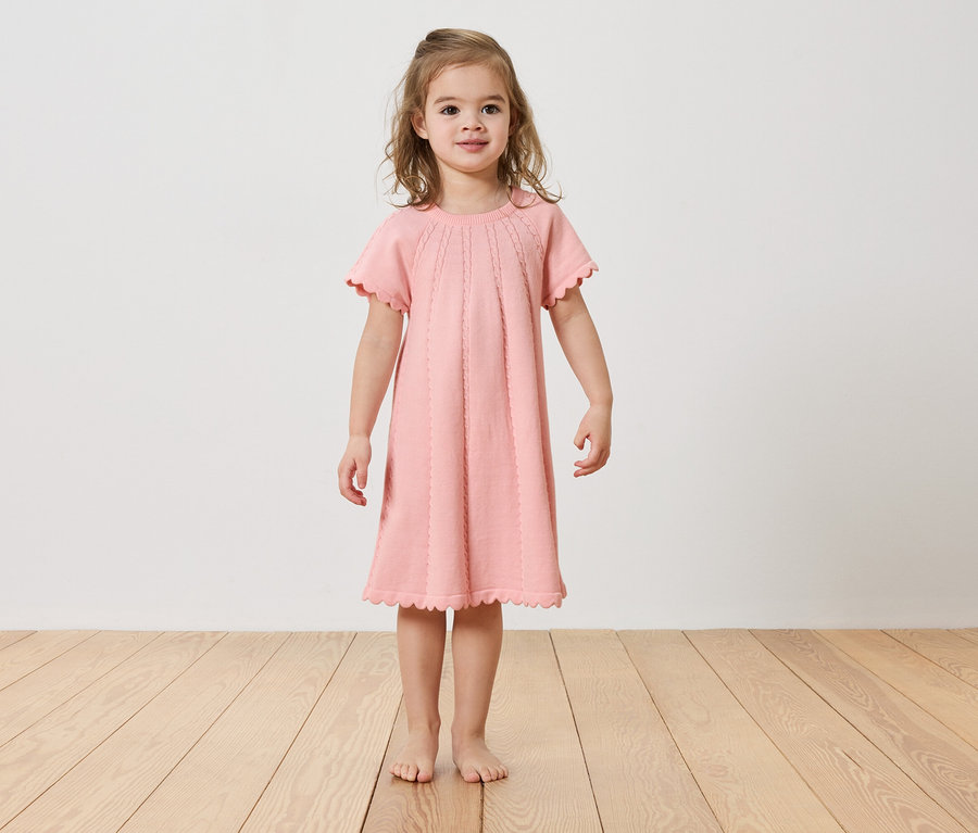 Mädchen steht barfuß auf einem Holzboden und trägt ein rosa Kleid.