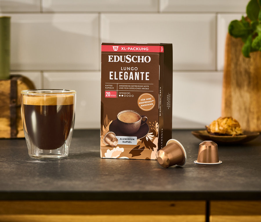 Ein Glas Kaffee steht neben einer Packung EDUSCHO Lungo Elegante - 20 Kapseln und zwei Kaffeekapseln auf einer Küchentheke.