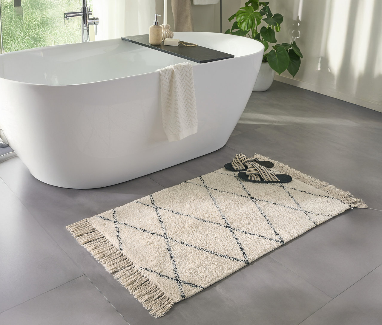 Ein Badezimmer mit einer weißen Badewanne und einer cremefarbenen benuta Badematte »Naomi« (50 x 80 cm) mit Fransen.
