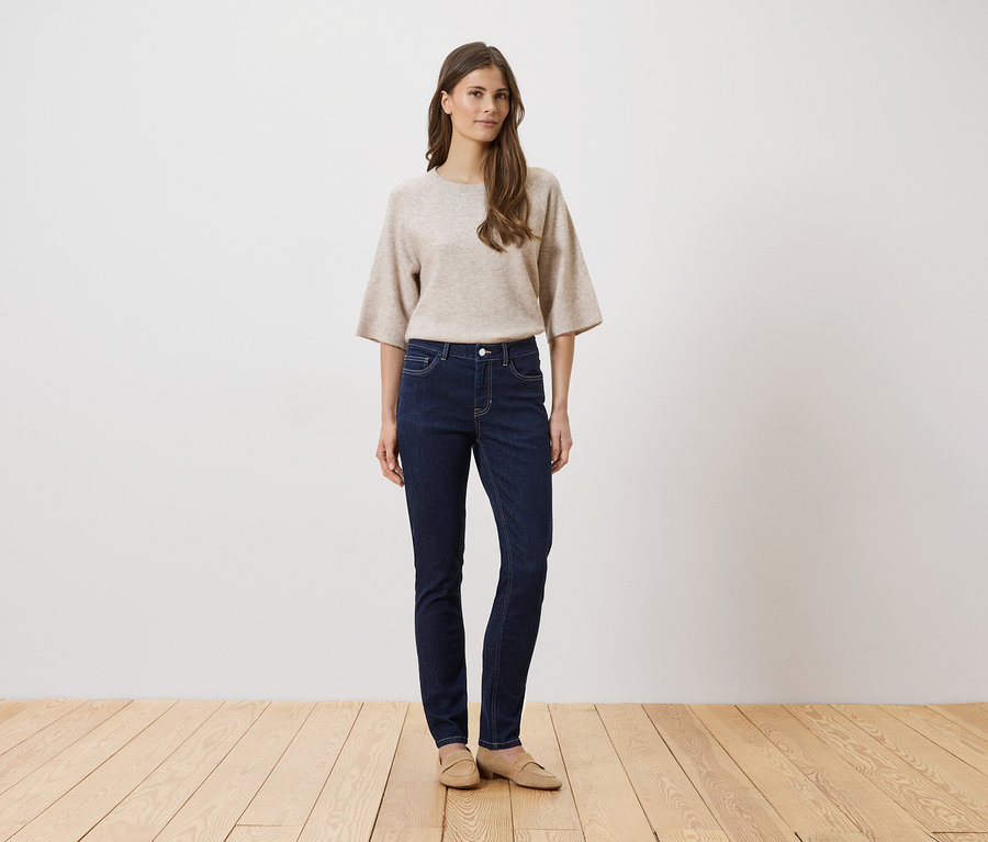 Frau mit beigem Pullover und dunkler Jeans steht auf einem Holzboden vor einer weißen Wand.