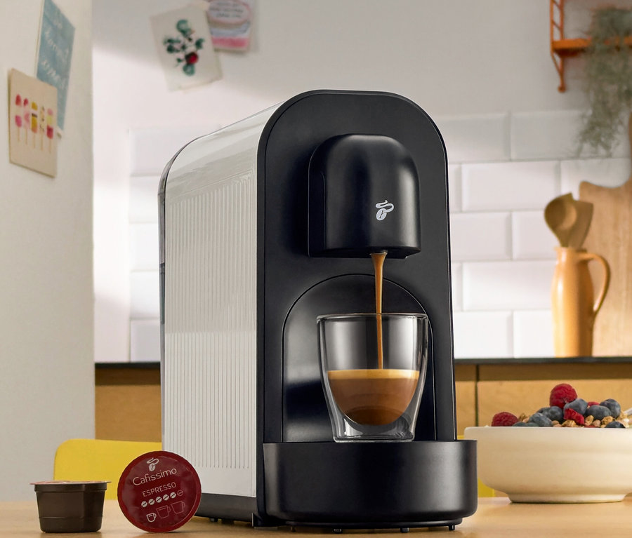 Eine schwarze Kaffeemaschine gießt Kaffee in ein Glas. Daneben liegen Kaffeepads und eine Schale mit Beeren.