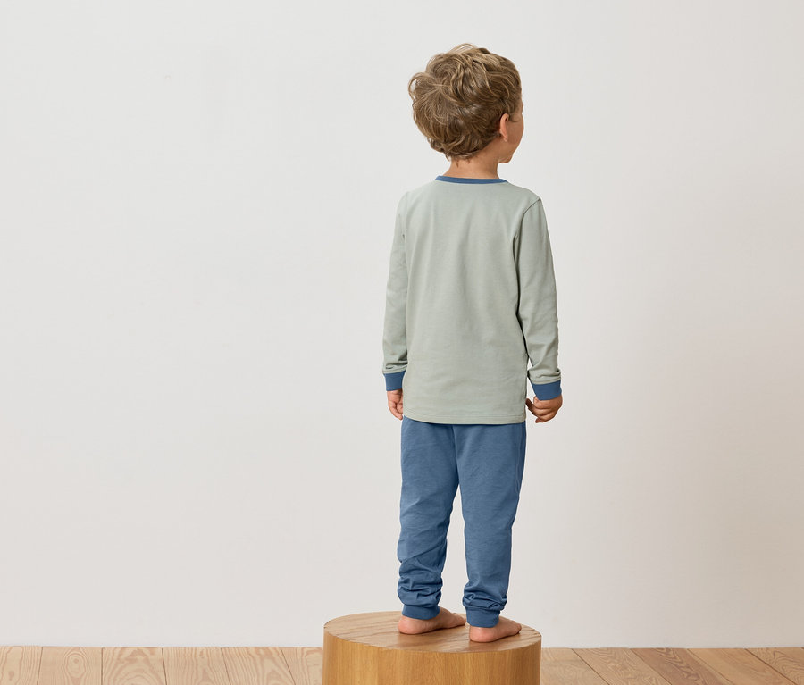 Ein Kind mit blonden Locken steht barfuß auf einem Holzhocker und trägt einen grünen Schlafanzug mit blauen Bündchen.