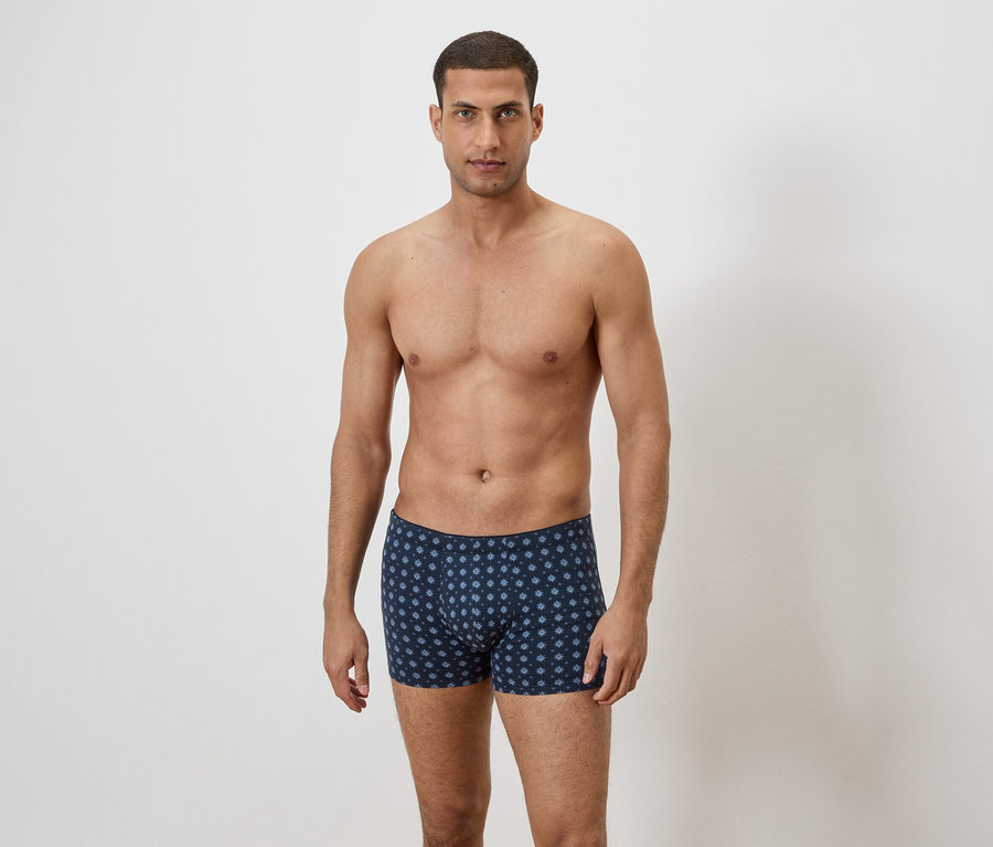 Ein Mann mit freiem Oberkörper steht vor einer weißen Wand und trägt blaue Boxershorts mit einem sich wiederholenden Blumenmuster.