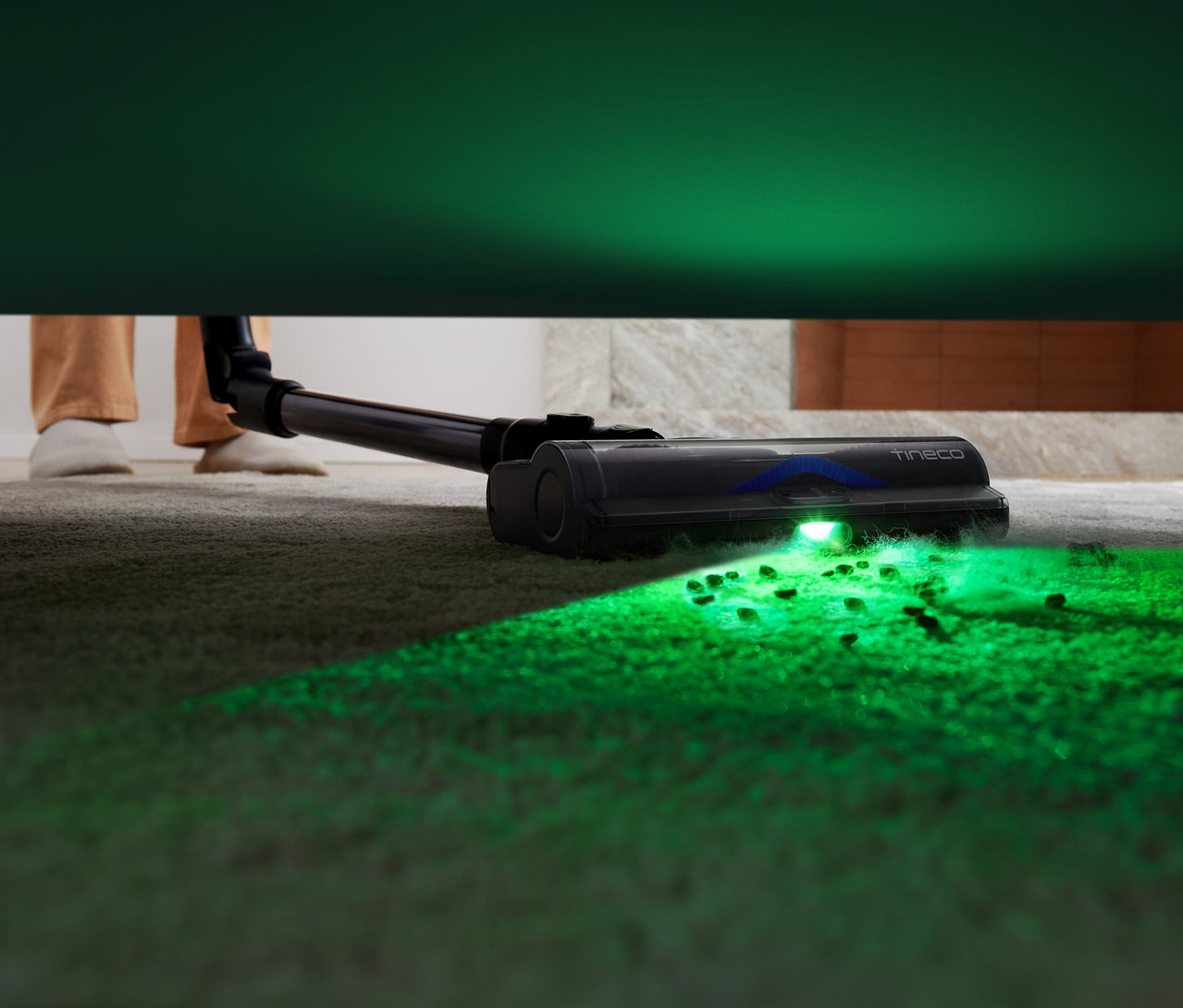 Eine Person saugt mit einem Tineco Akku-Staubsauger »Pure One S50 Pro« einen Teppich unter einem Sofa. Grünes Licht beleuchtet den Schmutz.