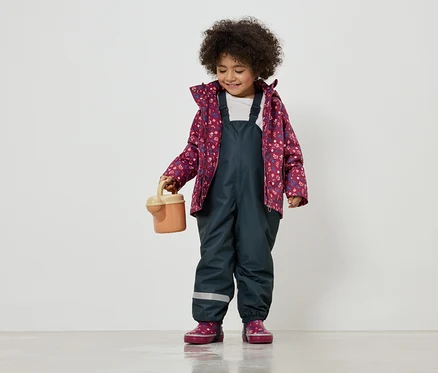 Ein Mädchen mit lockigem Haar trägt eine Blumenjacke, eine dunkelblaue Latzhose und hält eine kleine Gießkanne.