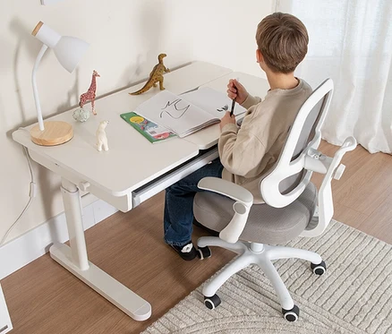 Junge sitzt an einem weißen Schreibtisch mit Lampe und Spielzeug und zeichnet in ein offenes Buch.
