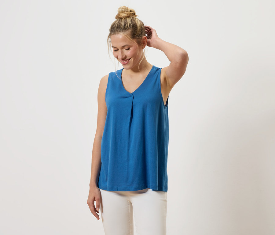 Frau mit blondem Haar trägt ein blaues V-Ausschnitt Top und weiße Hosen.