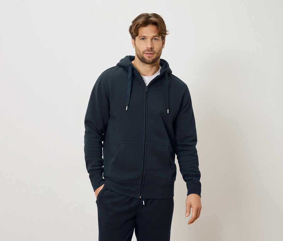 Mann mit marineblauem Reißverschluss-Hoodie und marineblauer Jogginghose, stehend vor einem cremefarbenen Hintergrund.