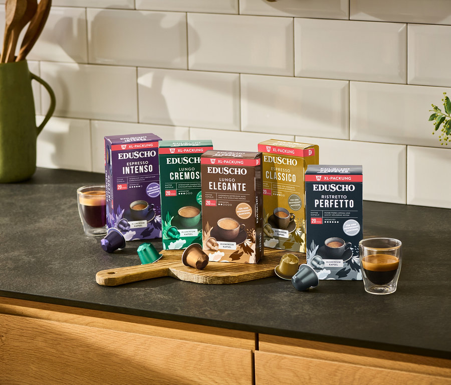 Fünf Packungen Eduscho Kaffeekapseln (Espresso Intenso, Lungo Cremoso, Lungo Elegante, Espresso Classico, Ristretto Perfetto) stehen auf einer Küchentheke mit zwei Tassen Kaffee und losen Kapseln.