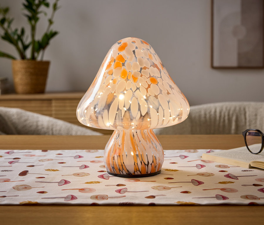 Eine pilzförmige Lampe aus orange-weiß gesprenkeltem Glas mit inneren Lichtern steht auf einem Holztisch. Darunter liegt ein weißer Tischläufer mit Pilzmustern. Rechts daneben liegen ein offenes Buch und eine Brille. Im unscharfen Hintergrund sind eine Topfpflanze und ein gerahmtes Bild zu sehen.