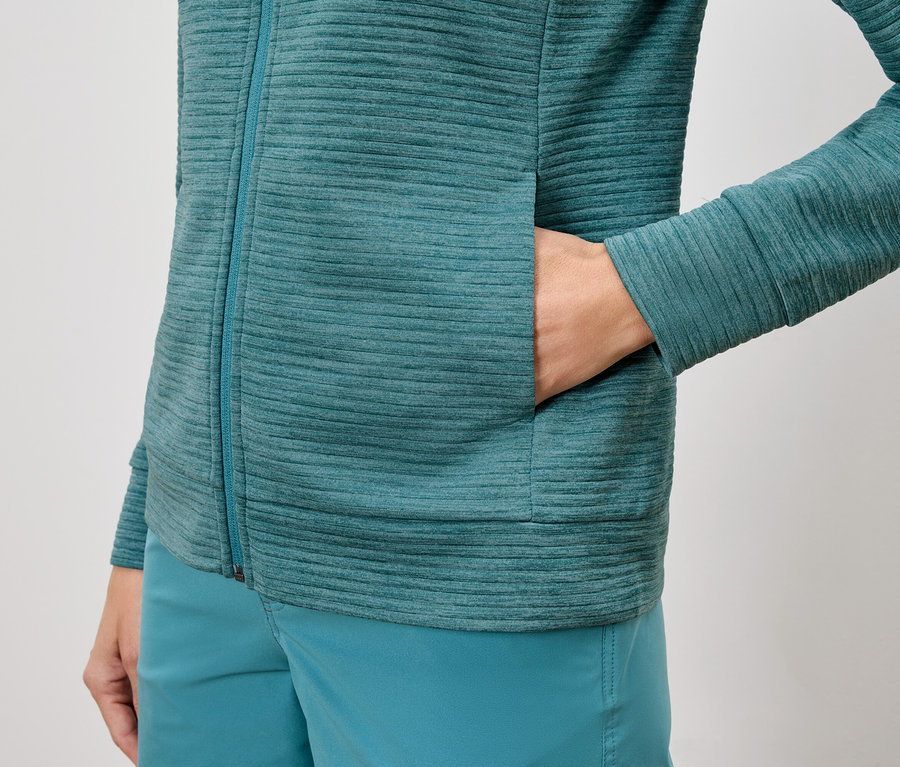 Nahaufnahme einer Person, die eine türkisfarbene Texturjacke und Shorts trägt, wobei die Hand in der Jackentasche steckt.