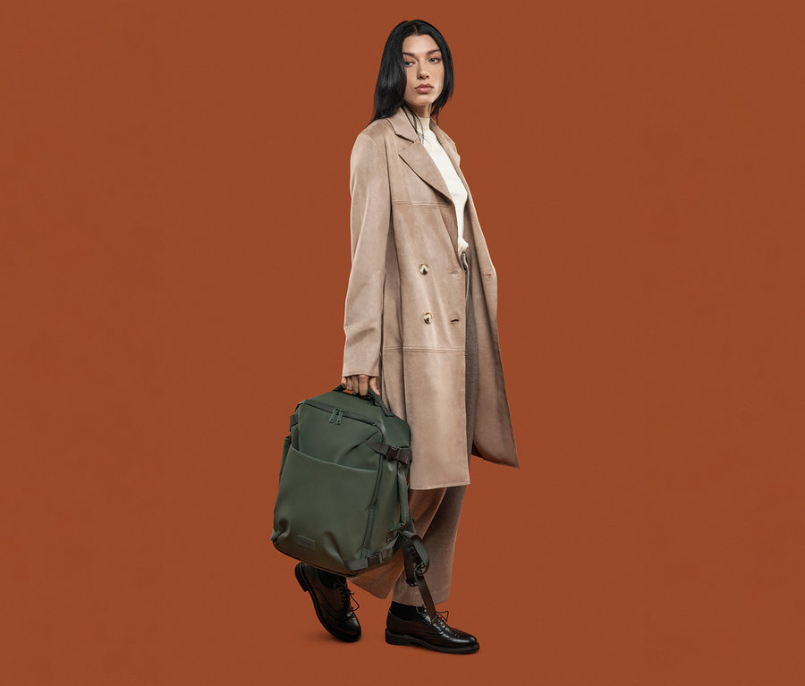 Frau mit beigem Mantel hält einen grünen Tucano TUGO DRY Kabinenrucksack mit 15,6"-Laptopfach.