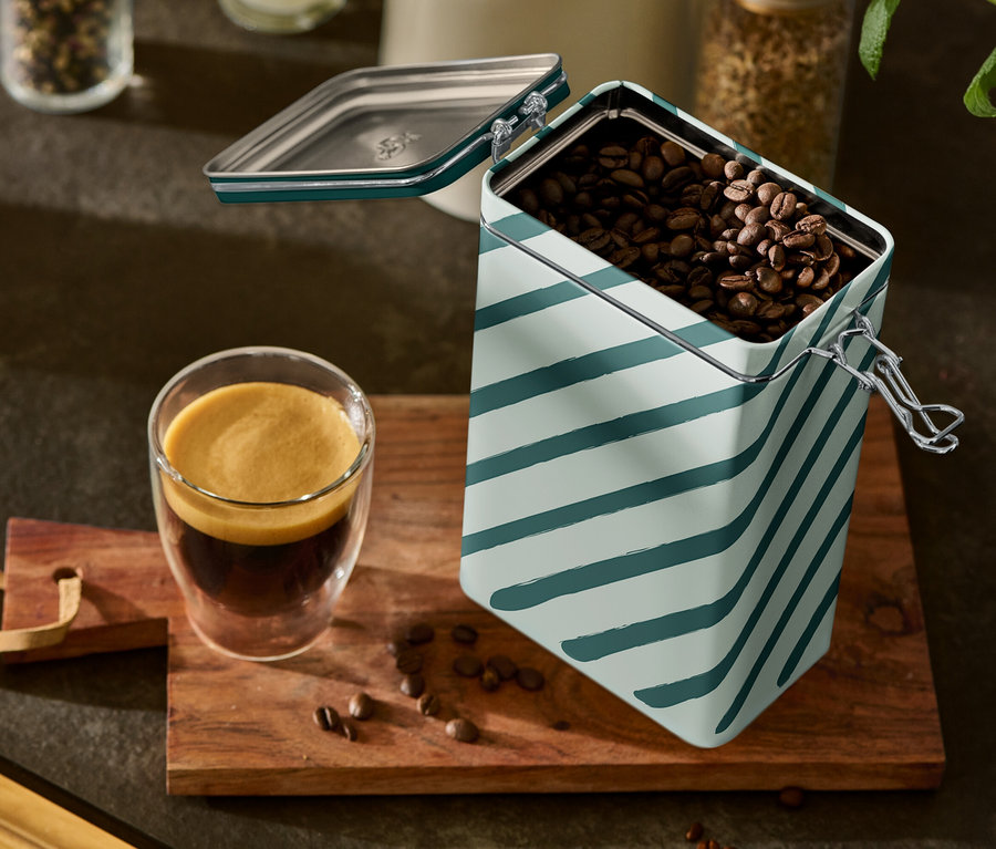 Ein Glas Espresso und eine geöffnete, gestreifte Kaffeedose stehen auf einem Holzbrett.