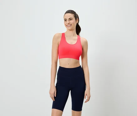 Frau mit Pferdeschwanz trägt einen pinken Sport-BH und kurze, marineblaue Leggings vor einem weißen Hintergrund.