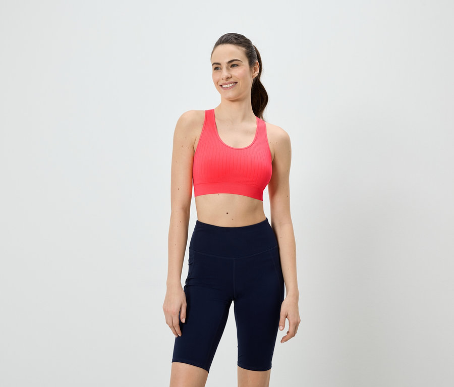 Frau mit Pferdeschwanz trägt einen pinken Sport-BH und kurze, marineblaue Leggings vor einem weißen Hintergrund.