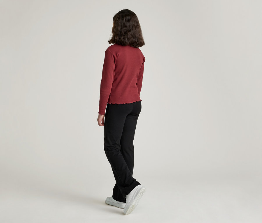 Eine Frau mit dunklen, schulterlangen Haaren steht mit dem Rücken zur Kamera und trägt ein weinrotes, langärmliges Oberteil, eine schwarze Hose und Turnschuhe.