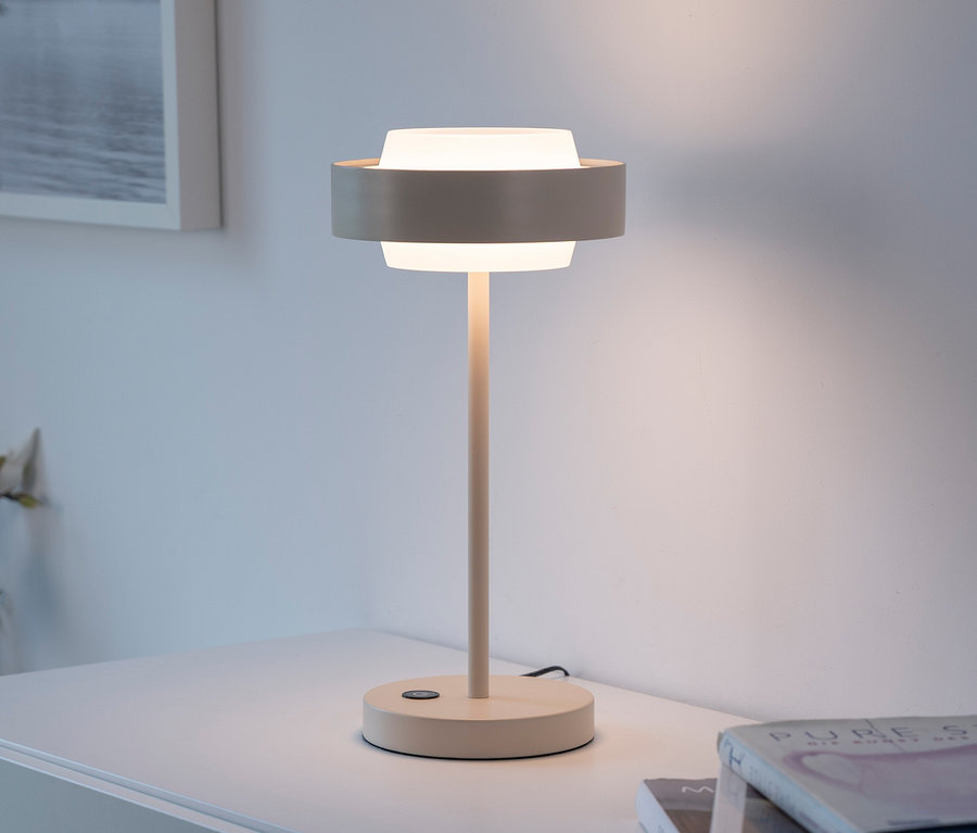 Eine leuchtende beigefarbene Tischlampe mit zylindrischem weißem Schirm und einem Ring in der Mitte steht auf einer weißen Kommode neben einem Stapel Bücher.