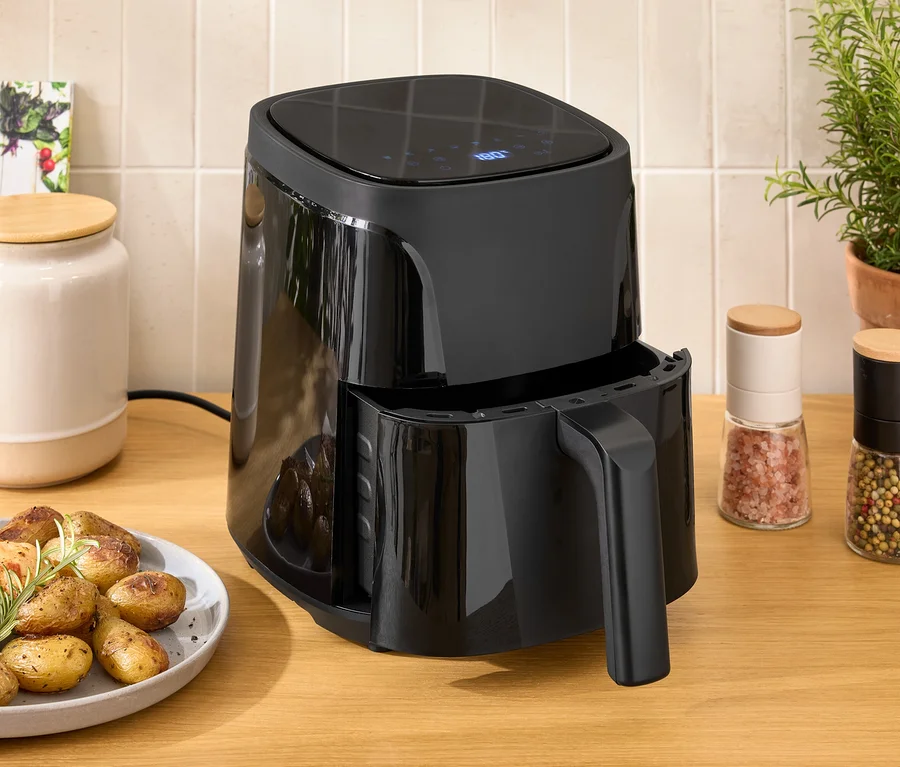 Eine schwarze Heißluftfritteuse steht auf einem Holztisch neben einem Teller mit Hühnchen und Kartoffeln.