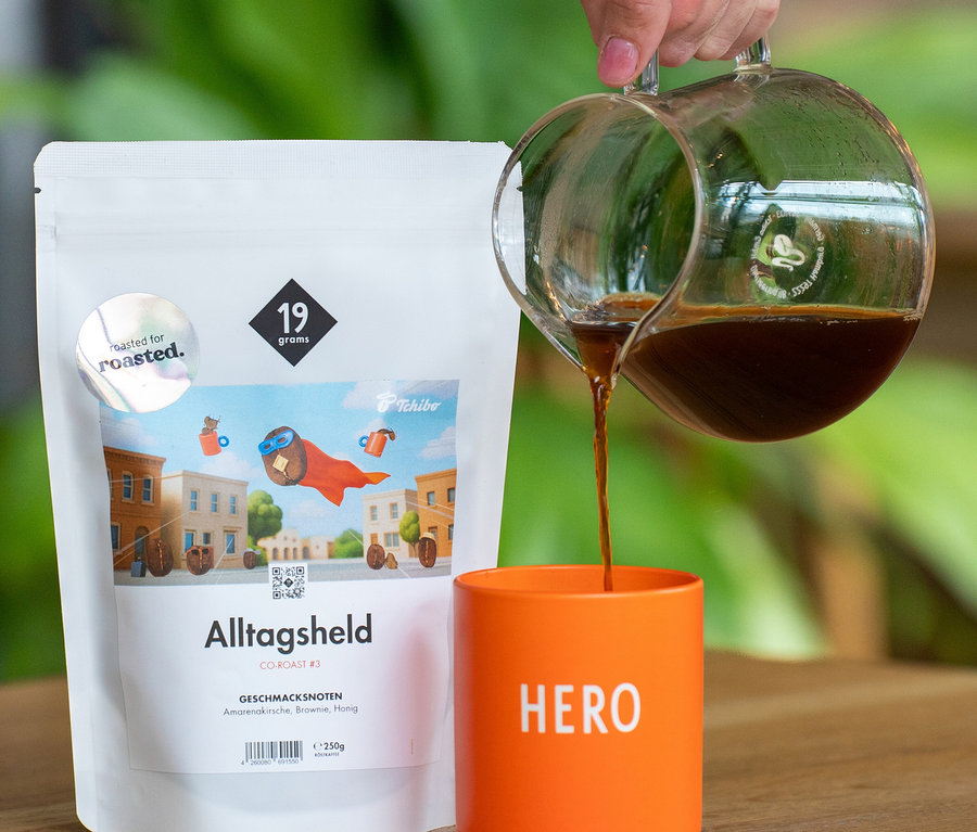 Eine Hand gießt Kaffee aus einer Glaskaraffe in einen orangefarbenen Becher mit „HERO“-Aufschrift. Daneben steht eine weiße 250g-Packung 19grams x roasted. - Alltagsheld Co-Roast #3 Omni-Roast Kaffeebohnen.