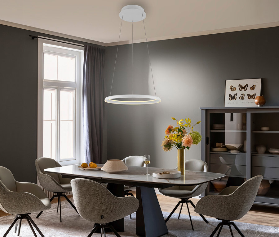 Ein modernes Esszimmer mit dunkelgrauen Wänden, einem rechteckigen Tisch mit Marmorplatte und sechs gepolsterten Stühlen. Darüber hängt die weiße SCHÖNER WOHNEN-Kollektion Pendelleuchte »Quillo«. Der Tisch ist mit bunten Blumen, Zitronen und Schalen dekoriert. Rechts steht ein grauer Vitrinenschrank mit Schmetterlingsbild.