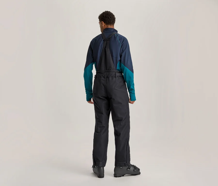 Mann von hinten in Skibekleidung, mit dunkelblau-türkisem Langarmshirt, schwarzer Skihose mit Trägern und schwarzen Skischuhen.