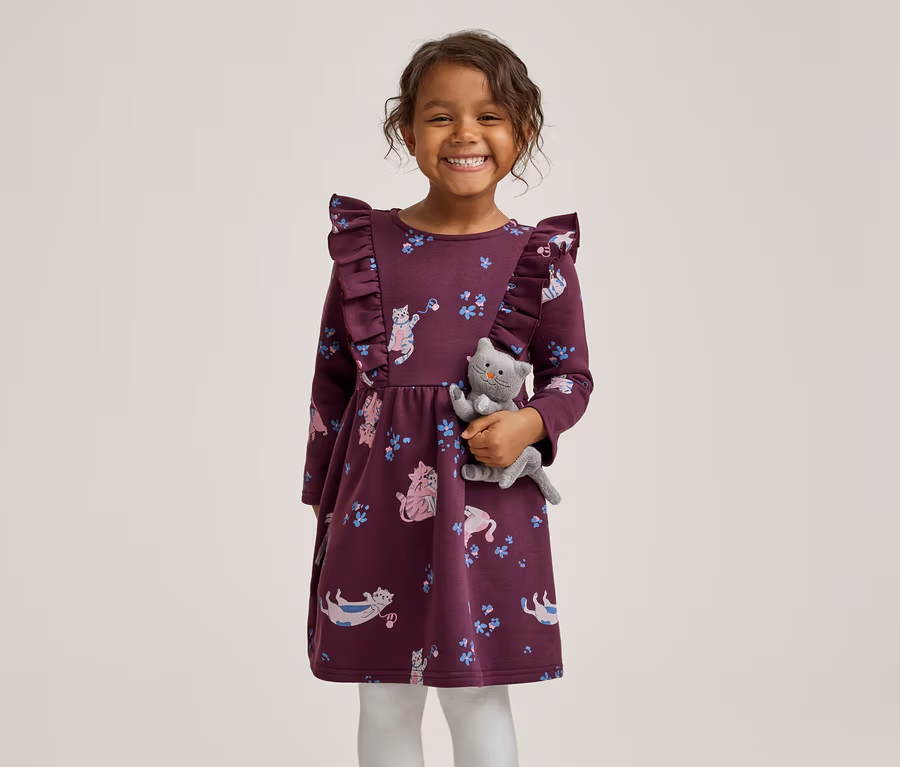 Ein lächelndes kleines Mädchen in einem lila Kleid mit Rüschen und Katzenmuster, das ein graues Plüschkätzchen hält.