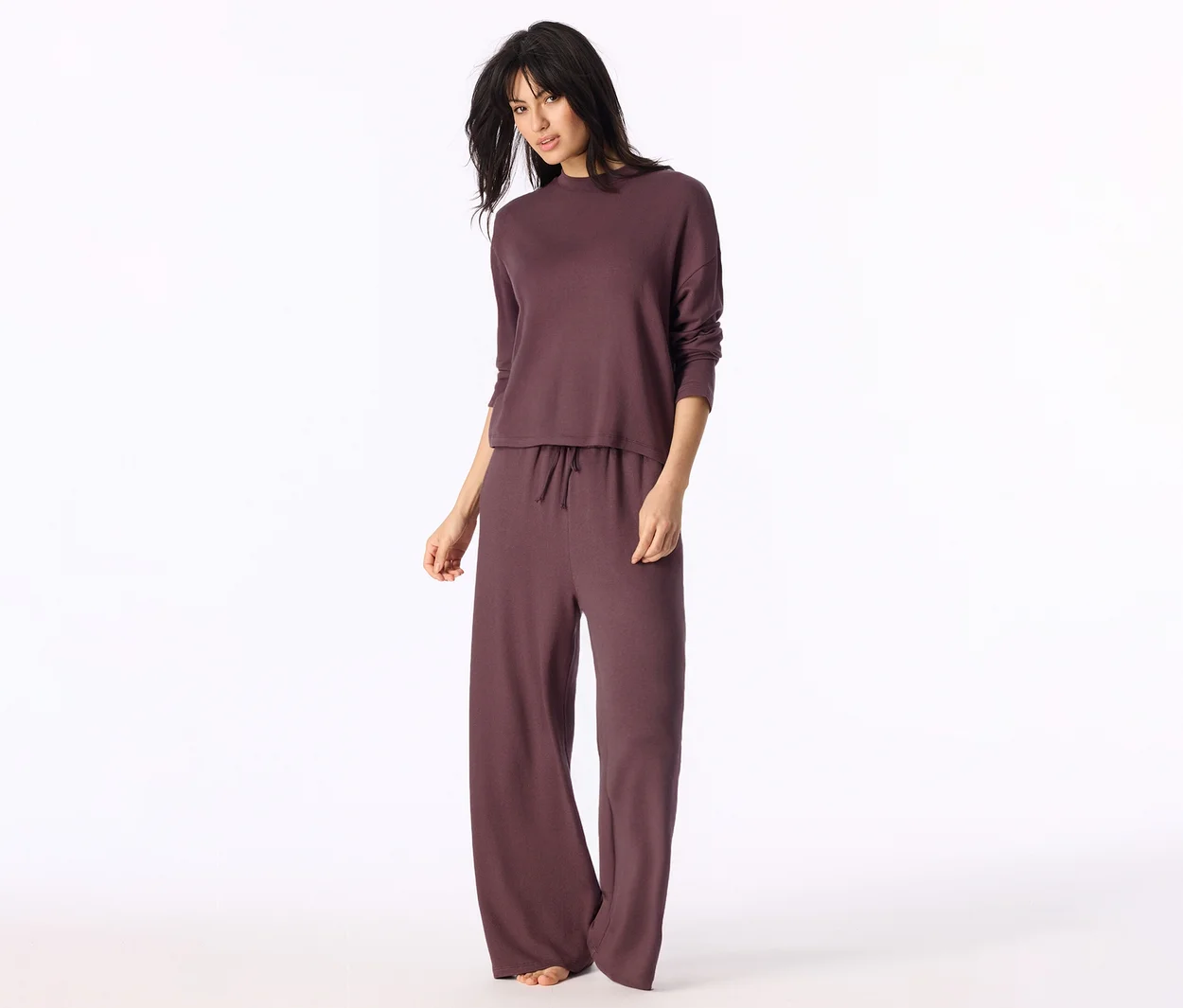 Frau in einem dunkelrotbraunen Loungewear-Set, bestehend aus langärmeligem Oberteil und weiter Hose mit Kordelzug. Sie steht barfuß und blickt in die Kamera.
