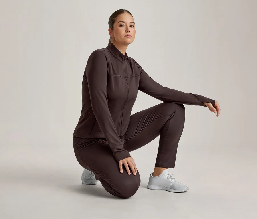 Frau in dunkelbrauner Sportjacke und passender Hose, hockend und in die Kamera blickend. Sie trägt hellgraue Sportschuhe.