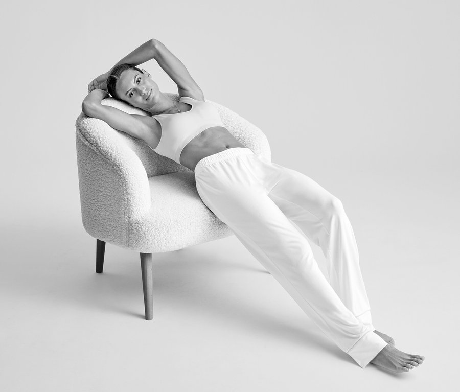Frau in einem cremefarbenen Seamless-Bustier und heller Hose lehnt sich mit den Armen hinter dem Kopf auf einem strukturierten Sessel zurück.
