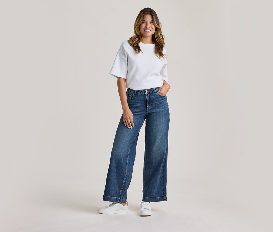 Lächelnde Frau steht, trägt ein weißes T-Shirt, weite blaue Jeans und weiße Sneaker, eine Hand in der Hosentasche.