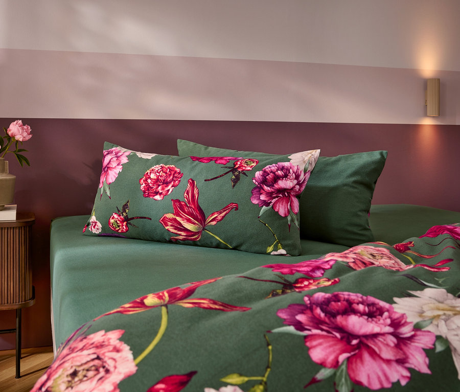 Ein Bett mit dunkelgrüner Bettwäsche, die mit großen rosa und roten Blumen gemustert ist, und einem einfarbigen grünen Kissen. Links steht ein Nachttisch mit einer Vase voller rosa Pfingstrosen. Die Wand ist zweifarbig in Rosa und Dunkelrot gestrichen.