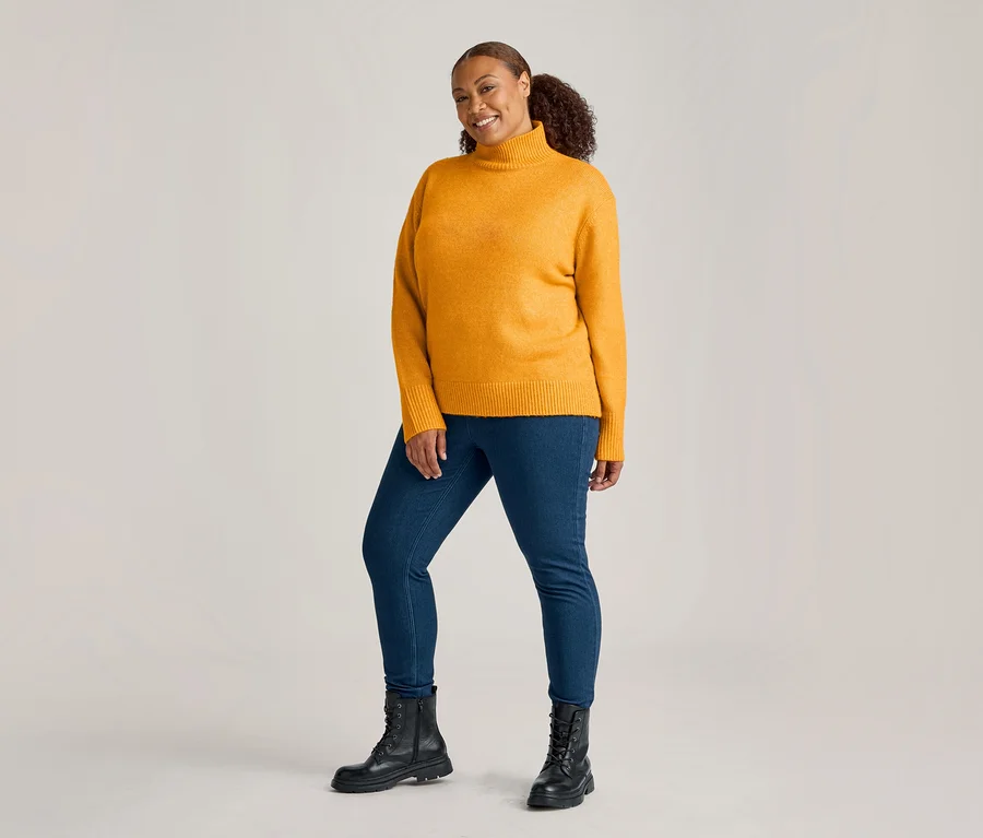 Frau in senfgelbem Rollkragenpullover, midblue Denim Jeggings und schwarzen Schnürstiefeln lächelt in die Kamera.