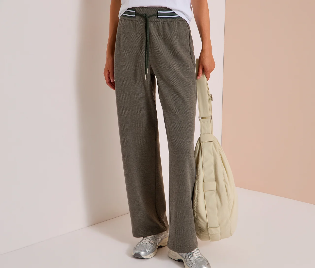 Eine Person trägt ein weißes T-Shirt, eine dunkelolivfarbene CECIL Stoffhose mit weitem Bein, gestreiftem Gummibund und Kordelzug sowie silberne Sneaker. Sie hält eine beigefarbene Tasche.