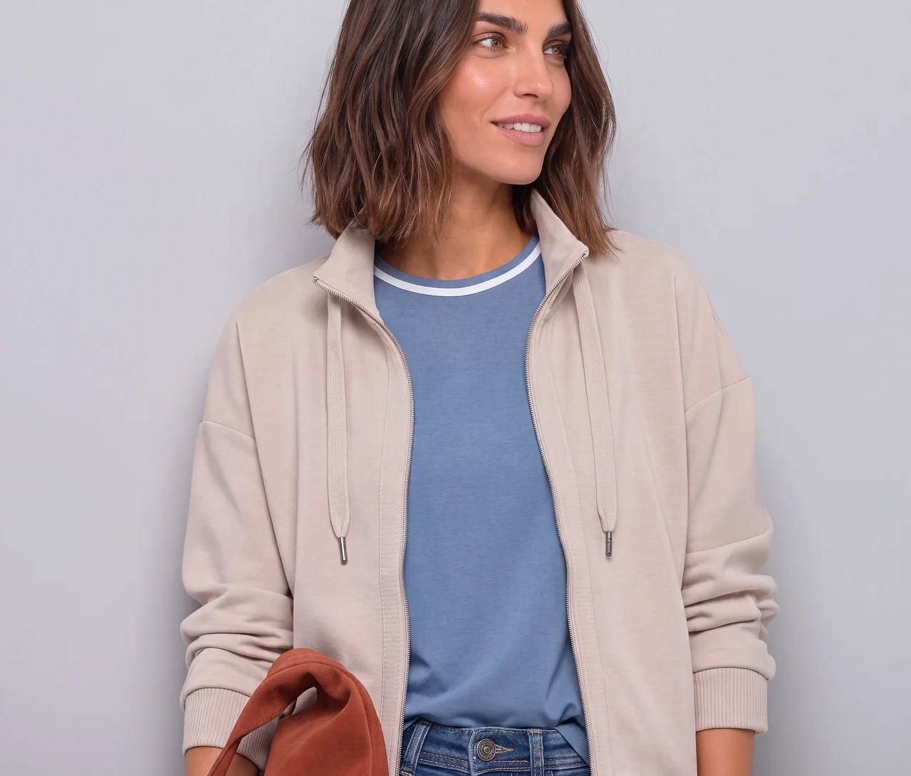 Eine Frau trägt eine offene beigefarbene Street One Sweatjacke über einem blauen T-Shirt und Jeans, während sie eine braune Tasche hält.