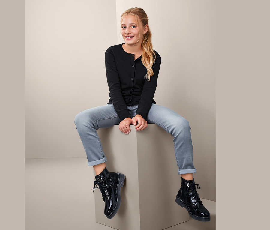 Mädchen sitzt auf einem Würfel und trägt eine Feinstrickjacke, Jeans und schwarze Stiefel.