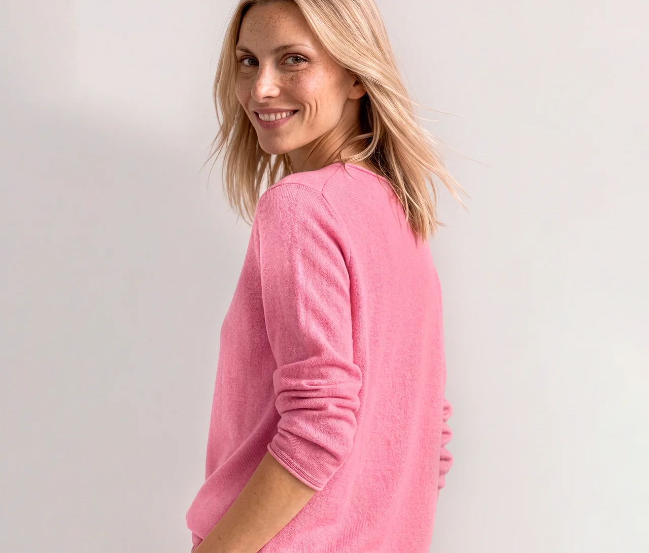 Eine lächelnde Frau mit blonden Haaren und Sommersprossen blickt über ihre Schulter und trägt einen roséfarbenen Street One Strickpullover.
