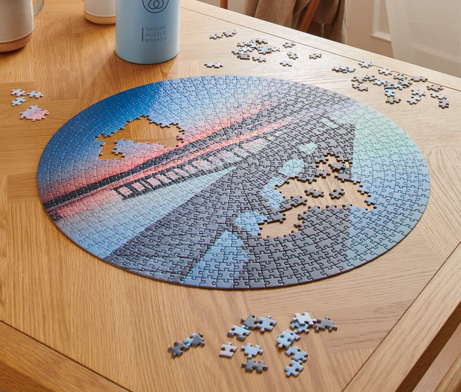Auf einem Holztisch liegt ein rundes Puzzle »Steg« mit einem Brückenmotiv, das teilweise zusammengesetzt ist, und daneben liegen Puzzleteile.