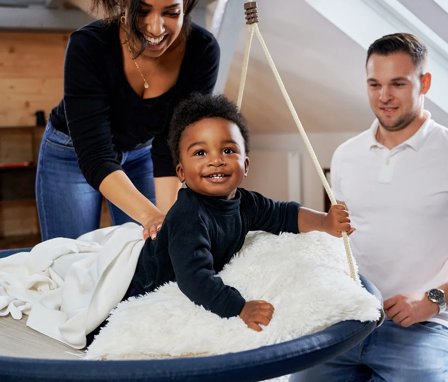 Ein Kind sitzt in einer schwarzen HUDORA Nestschaukel »Lounge« mit 90 cm Durchmesser, während die Mutter dahinter steht und der Vater daneben kniet.