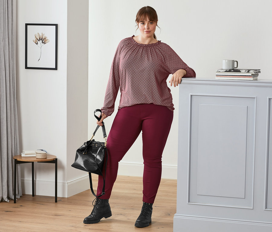 Frau posiert mit dunkelrotem, bedrucktem Blusenshirt, dunkelroter Stretchhose, Leder-Boots und Beuteltasche.