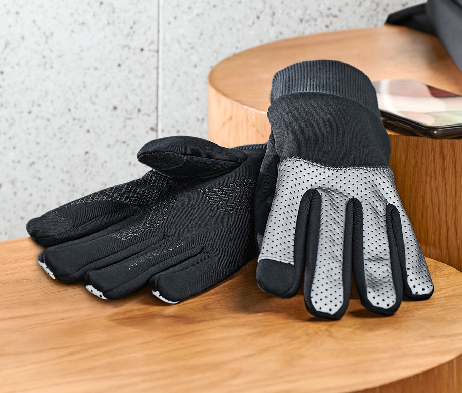 Schwarze Windprotection-Handschuhe mit Reflektorbesatz liegen auf einem Holztisch neben einem Handy und einer schwarzen Jacke.