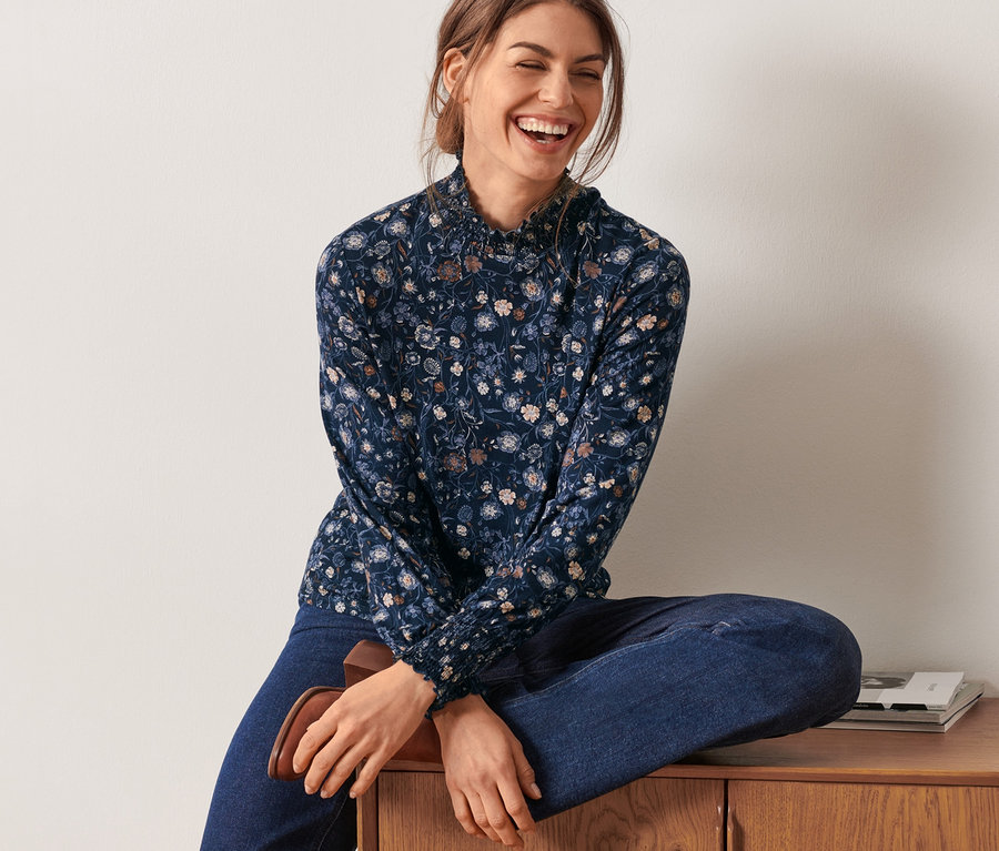 Frau sitzt auf einer Holzkommode und trägt ein florales Blusenshirt mit Stehkragen.