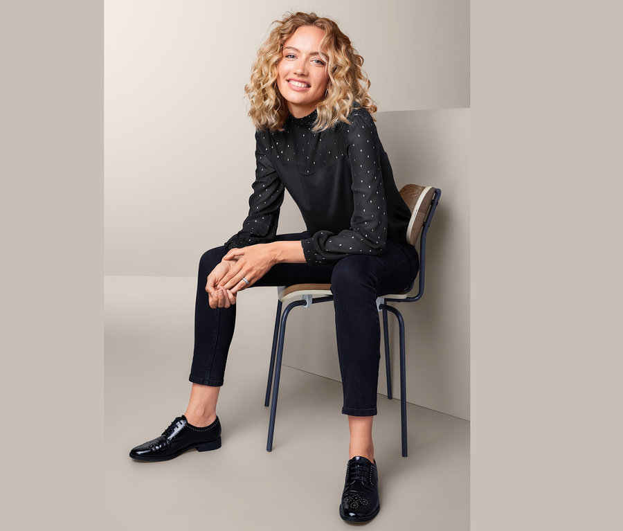Frau sitzt auf einem Stuhl und trägt ein Blusenshirt im Materialmix, schwarze Hose und schwarze Schuhe.