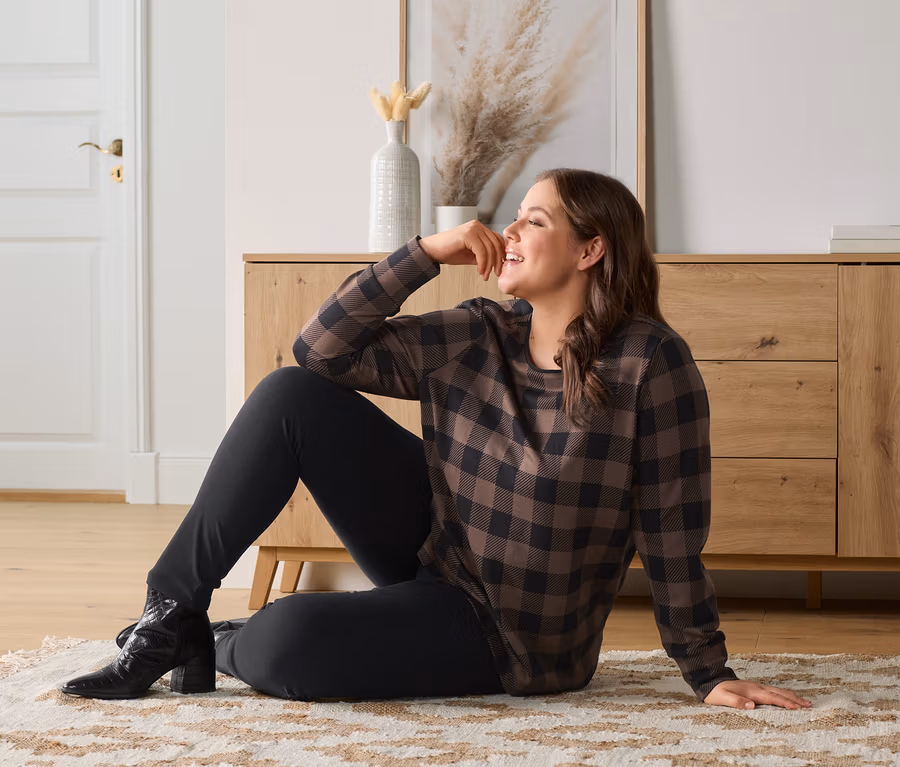 Eine Frau sitzt auf dem Teppichboden und trägt ein kariertes Sweatshirt, Jeggings und geprägte Leder-Stiefeletten.