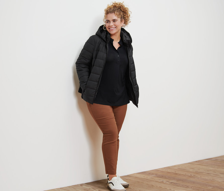 Frau posiert in schwarzer Steppjacke mit Kapuze, Blusenshirt mit Knopfleiste, cognacfarbener Stretchhose und Lederboots.